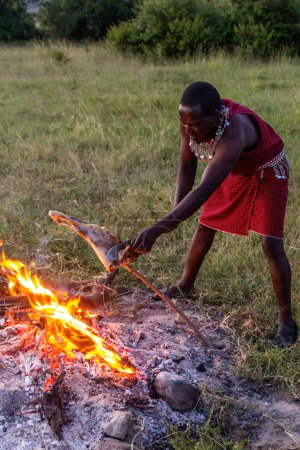 Foto de MASAI MARA, KENIA - 20 DE FEBRERO DE 2020: Masai hombre asando una pierna de cabra, Kenia - Imagen libre de derechos