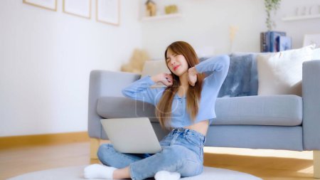 Foto de Concepto de síndrome de oficina. Mujer joven asiática que trabaja en el ordenador portátil mientras está sentada en el suelo junto al sofá en casa, sintiendo dolor en el cuello y el hombro después de trabajar en el ordenador portátil durante mucho tiempo - Imagen libre de derechos