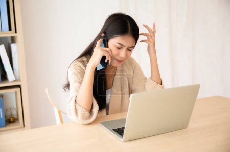 Téléchargez les photos : Jeune femme asiatique parlant smartphone mobile tout en travaillant sur ordinateur portable à la maison. Travail à domicile, vidéoconférence, vidéoconférence - en image libre de droit