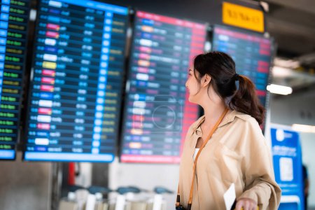 Téléchargez les photos : Jeune femme asiatique avec passeport et carte d'embarquement en main à l'aéroport international en regardant le tableau d'information de vol, vérifier son vol - en image libre de droit