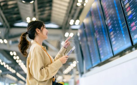Young asian woman with passport and boarding pass as a hand in international airport looking at the flight information board, checking her flight