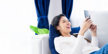 Téléchargez les photos : Happy young asian woman relaxing at home. Female smile lying on sofa and holding mobile smartphone. Girl using video call to friend - en image libre de droit
