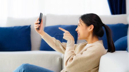 Téléchargez les photos : Happy young asian woman relaxing at home. Female smile sitting beside sofa and holding mobile smartphone. Girl using video call to friend - en image libre de droit