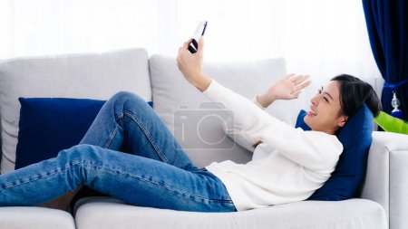 Téléchargez les photos : Happy young asian woman relaxing at home. Female smile lying on sofa and holding mobile smartphone. Girl using video call to friend - en image libre de droit