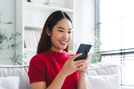 Photo for Happy young asian woman relax on comfortable couch at home texting messaging on smartphone, smiling girl use cellphone chatting, online shopping at home, video call communication - Royalty Free Image