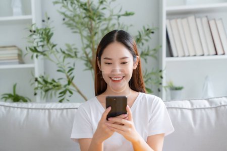 Photo for Happy young Asian woman relax sitting on couch using cell phone, smiling lady laughing holding smartphone, looking at cellphone enjoying doing online ecommerce shopping in mobile apps - Royalty Free Image