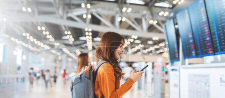 Photo for Young asian woman in international airport, using mobile smartphone and checking flight at the flight information board - Royalty Free Image
