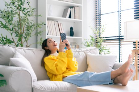 Photo for Happy young asian woman  wearing headset and relaxing on comfortable couch at home, texting messaging on smartphone, smiling girl use cellphone chatting, online shopping at home, video call - Royalty Free Image