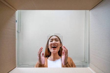 Photo for Low angle view of surprised young asian woman unpacking. Opening carton box and looking inside. Packaging box, delivery service. Human emotions and facial expression - Royalty Free Image