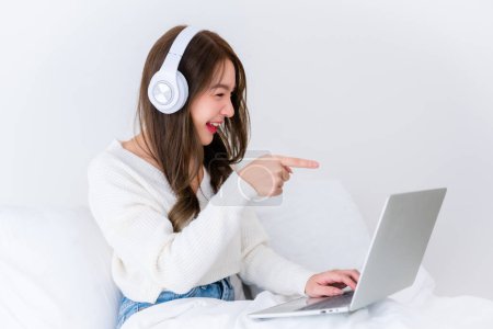 Photo for Morning Time in Bedroom Young Asian Woman Happy Working on Computer Laptop, Wearing headset, Sitting on Bed at House - Royalty Free Image