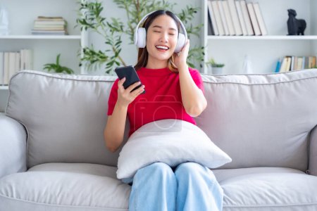 Photo for Young Asian woman relaxing on sofa at home, listening music with headset on cell phone. People lifestyle concept - Royalty Free Image