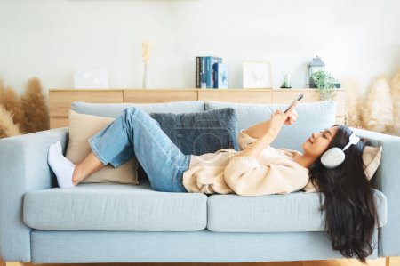 Photo for Happy young asian woman relaxing at home. Female smile sitting on sofa and holding mobile smartphone. Girl using video call to friend. Listening to music - Royalty Free Image