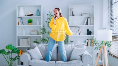 Photo for On the sofa in her living room, a young Asian woman dances. Asian woman with a smile relaxing at home, happy and in good mental and physical health, wearing headset and listening to music - Royalty Free Image