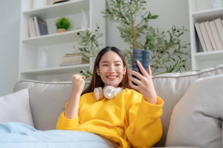 Photo for Happy young asian woman relax on comfortable couch at home texting messaging on smartphone, smiling girl use cellphone, chatting online message, shopping online from home - Royalty Free Image