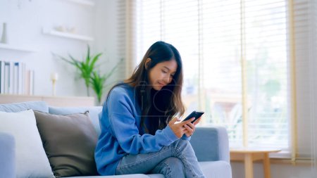 Photo for Happy young asian woman relax on comfortable couch at home texting messaging on smartphone, smiling girl use cellphone chatting, browse wireless internet on gadget, shopping online from home - Royalty Free Image