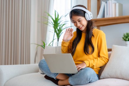 Photo for Young adorable asian woman smile wearing headset working in living room at home. Happy gorgeous female using computer laptop and sitting on couch at house, Video call with friend - Royalty Free Image