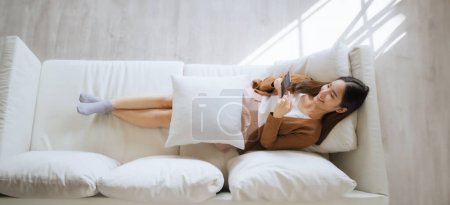 Photo for High angle view of happy cheerful positive young Asian woman on the phone at home, smiling girl using app and browsing on the internet on smartphone lying on sofa - Royalty Free Image