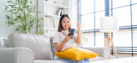 Photo for Happy young asian woman relax on comfortable couch at home texting messaging on smartphone, smiling girl use cellphone, chatting online message, shopping online from home - Royalty Free Image