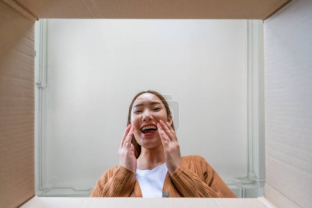 Photo for Low angle view of surprised young asian woman unpacking. Opening carton box and looking inside. Packaging box, delivery service. Human emotions and facial expression - Royalty Free Image