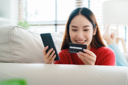 Photo for Young asian woman lying on sofa in living room, makes online banking payments through the internet from bank card on cell phone. Shopping online on mobile with credit card - Royalty Free Image