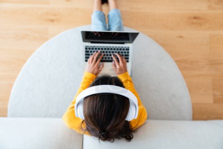 Photo for Top view of young Asian woman sitting on the floor and working on computer laptop, wearing wireless headset, listening to music - Royalty Free Image