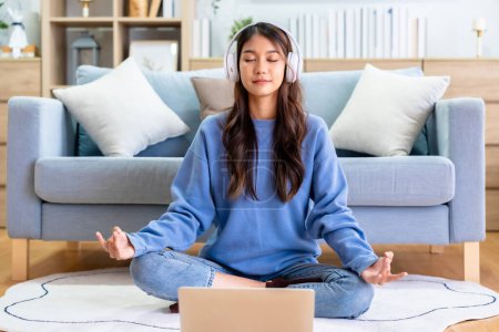 Photo for Happy young Asian woman practicing yoga and meditation at home sitting on floor in living room in lotus position and relaxing with closed eyes. Mindful meditation and wellbeing concept - Royalty Free Image