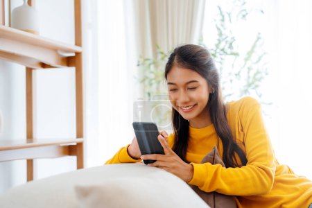 Photo for Happy young Asian woman sitting on sofa holding mobile phone using cellphone technology doing ecommerce shopping, buying online, texting messages relaxing on couch in cozy living room at home - Royalty Free Image