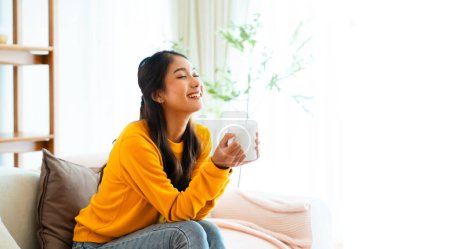 Photo for Beauty young Asian woman enjoying a cup of coffee at home. Smiling pretty girl drinking hot milk and sitting on couch - Royalty Free Image