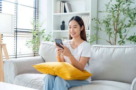 Photo for Happy young asian woman relax on comfortable couch at home texting messaging on smartphone, smiling girl use cellphone chatting, online shopping at home, video call communication - Royalty Free Image