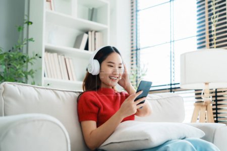 Photo for Young Asian woman relaxing on sofa at home, listening music with headset on cell phone. People lifestyle concept - Royalty Free Image
