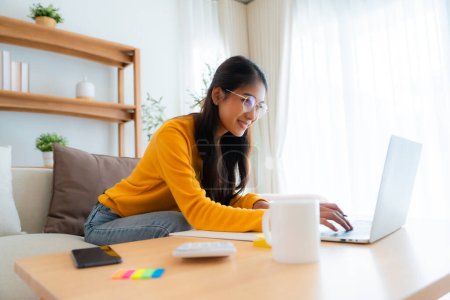 Photo for Young Asian female wearing glasses using laptop, working at home in living room, coffee mug on table. Cozy office workplace, remote work - Royalty Free Image