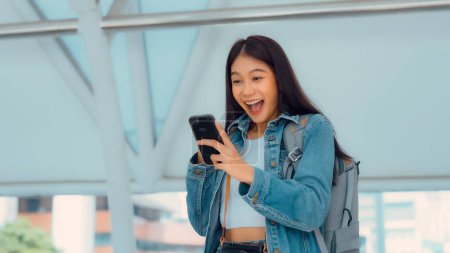 Photo for Young asian woman using mobile in city. Happy female tourist wearing jeans jacket and holding smartphone at public - Royalty Free Image
