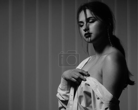 Téléchargez les photos : Noir et blanc photo d'une belle jeune fille dans une seule chemise blanche sur son corps nu posant dans un studio - en image libre de droit