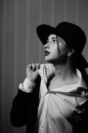 Téléchargez les photos : Photo noir et blanc d'une belle jeune fille dans un seul blazer noir et chapeau posant dans un studio - en image libre de droit
