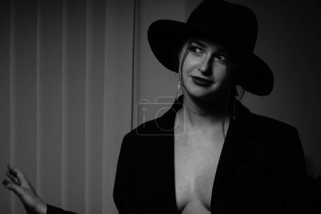 Téléchargez les photos : Photo noir et blanc d'une belle jeune fille dans un seul blazer noir sur son corps nu et chapeau posant dans un studio - en image libre de droit