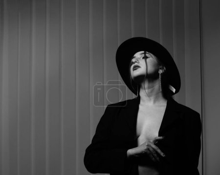 Foto de Foto en blanco y negro de una hermosa joven en una sola chaqueta negra sobre su cuerpo desnudo y sombrero posando en un estudio - Imagen libre de derechos