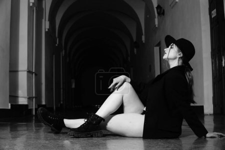 Téléchargez les photos : Photo noir et blanc d'une belle jeune fille dans un seul blazer noir sur son corps nu et chapeau posant dans un studio - en image libre de droit
