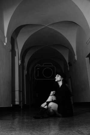 Foto de Foto en blanco y negro de una hermosa joven en una sola chaqueta negra sobre su cuerpo desnudo y sombrero posando en un estudio - Imagen libre de derechos