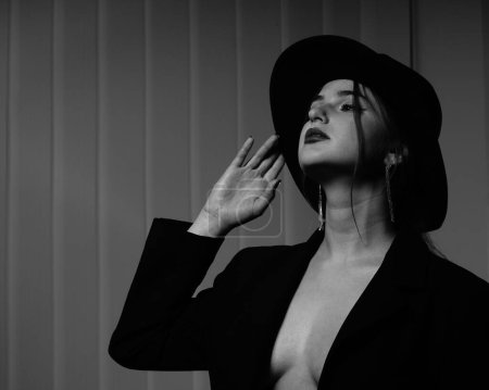 Photo for Black and white photo of a beautiful young girl in only one black blazer over her naked body and hat posing in a studio - Royalty Free Image