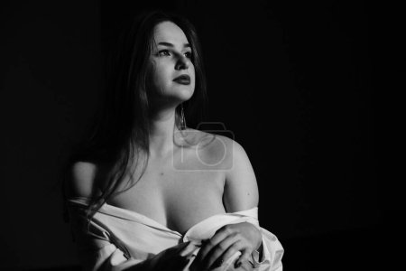 Téléchargez les photos : Noir et blanc photo d'une belle jeune fille dans une seule chemise blanche sur son corps nu posant dans un studio - en image libre de droit