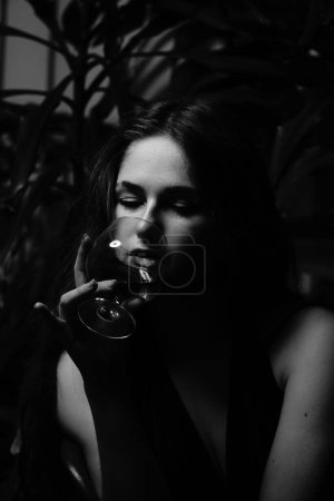 Téléchargez les photos : Photo noir et blanc d'une belle femme sensuelle dans une robe noire avec un verre de whisky posant pour l'appareil photo - en image libre de droit