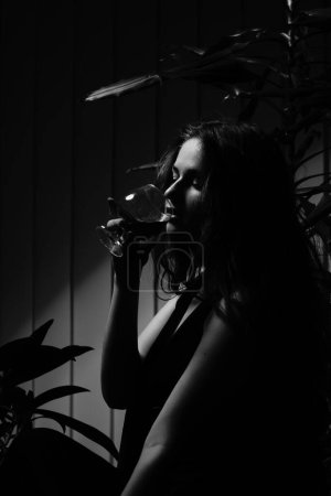 Photo for Black and white photo of a beautiful sensual woman in a black dress with a glass of whiskey posing for the camera - Royalty Free Image