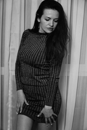 Foto de Foto en blanco y negro de una buena mujer en un vestido corto a rayas de pie cerca de las cortinas de la habitación - Imagen libre de derechos