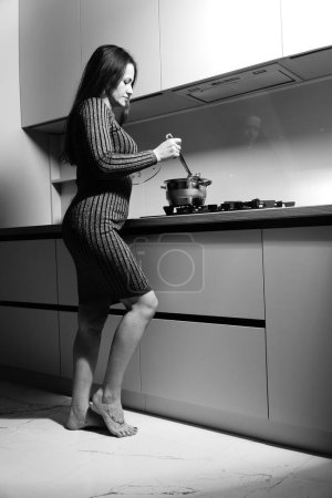 Foto de Foto en blanco y negro de una buena mujer en un vestido corto a rayas cerca de la estufa en la cocina - Imagen libre de derechos