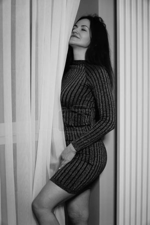 Foto de Foto en blanco y negro de una buena mujer en un vestido corto a rayas de pie cerca de las cortinas de la habitación - Imagen libre de derechos