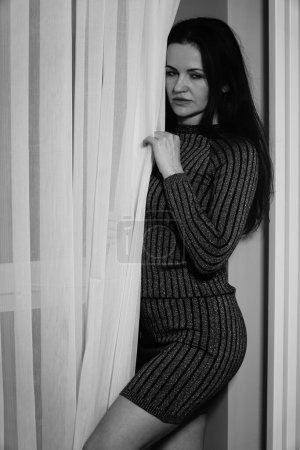 Téléchargez les photos : Photo noir et blanc d'une belle femme dans une robe courte rayée debout près des rideaux dans la chambre - en image libre de droit