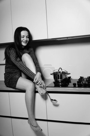Téléchargez les photos : Photo noir et blanc d'une jolie femme dans une robe courte rayée assise sur la table près du poêle dans la cuisine - en image libre de droit