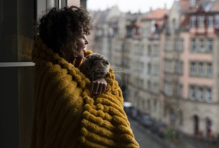  Frau mit lockigem Haar in Decke mit einer Katze, die bei kaltem Wetter am Fenster sitzt