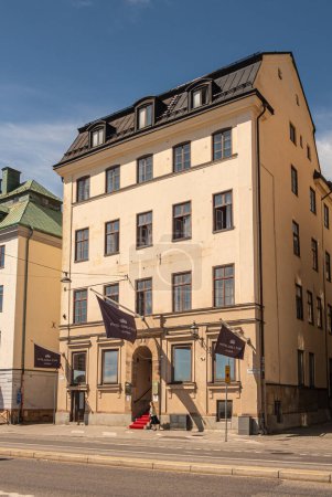 Sweden, Stockholm - July 17, 2022: Closeup of Hotel Gamla stan yellow classic building on Skeppsbron quay. Brown flags with name up front, red carpet, and pedestrian under blue sky