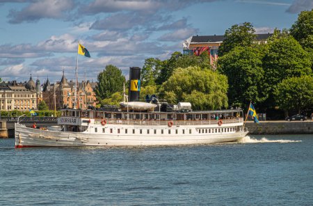Téléchargez les photos : Suède, Stockholm - 17 juillet 2022 : Bateau à vapeur Storskar blanc historique à partir de Skeppsbron quay. Ferry et tourizt voir l'entreprise sous un paysage nuageux bleu. Feuillage vert comme toile de fond - en image libre de droit